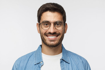 Young handsome student isolated on gray background, in blue shirt, wearing trendy round glasses, looking at camera with happy smile