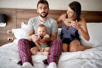 people, rest, love, relationships and television concept - happy couple and their baby son with remote lying in bed at home and watching tv.