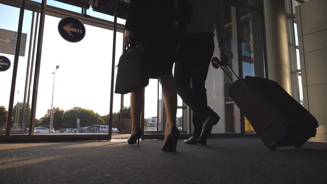 Businessman with his female colleague leaving terminal through glass doors and roll suitcase on wheels. Business man and woman going from the airport to city street with their luggage. Slow motion