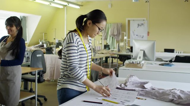 Medium shot of concentrated young Asian woman taking notes in sewing patterns and using swatches to choose color for clothes, her colleagues working in the background