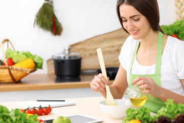 Beautiful Hispanic woman cooking in kitchen while using tablet computer and wooden spoon. Housewife found new recipe for dinner or breakfast. Healthy meal and householding concepts