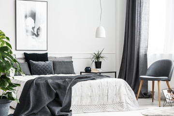 Modern, gray chair with wooden legs by a window of a bright bedroom interior with a dark gray blanket on a comfy bed
