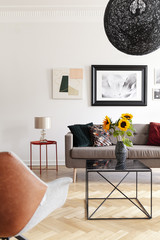 Sunflowers on table in modern living room interior with lamp and posters above sofa. Real photo