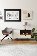 Grey armchair next to white cabinet with lamp in modern loft interior with posters. Real photo