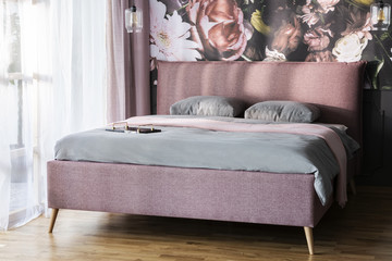 Pink blanket and grey cushions on bed in bright bedroom interior with flower print on the wall....