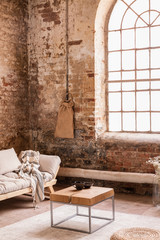 Blanket on grey couch in simple red brick loft interior with wooden table and window. Real photo