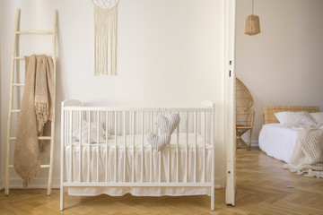 Fototapeta na wymiar Wooden crib with cushions standing in real photo of white kid room interior with blanket on ladder and macrame on the wall