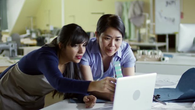 Medium shot of two multiethnic women using laptop computer and color swatches when choosing design ideas for fashion collection