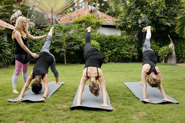 Group of young women practising one-legged downward facing dog pose under control of female yoga teacher outdoors