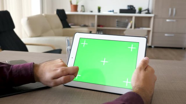 Man working on digital tablet PC with green screen mock up. The gadget is on the desk. In blurred background a comfortable living room. Dolly slider 4K footage with parallax effect