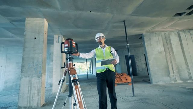 Male engineer is coming into a construction site and working with leveling equipment