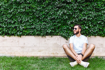 Stylish hipster with a beard and a white t-shirt, sitting on the lawn at the deciduous green wall