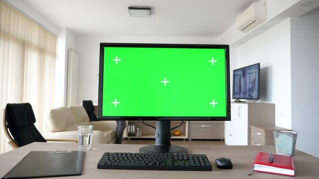 Personal PC computer with big green screen chroma mock up on the table in the living room. A guy is entering the room in background while the TV is on and sits on the sofa looking at his phone