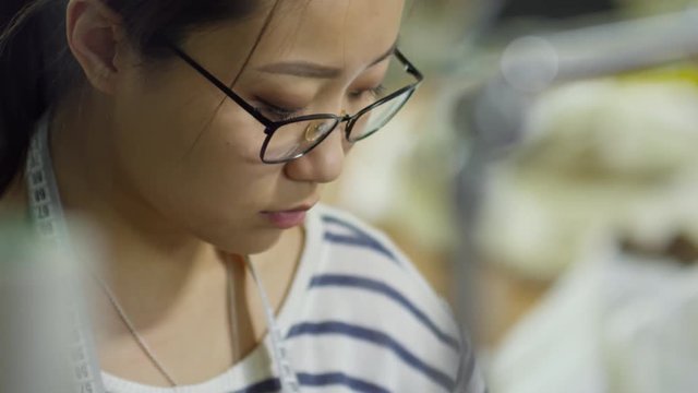 Close up face of focused young Asian seamstress in glasses working at sewing factory and making clothes