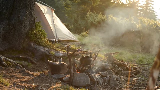 Old small kettle is heated on a bright bonfire on a green mountain meadow next to tent in the rays of rising sun - stunning morning during travel in the mountains.