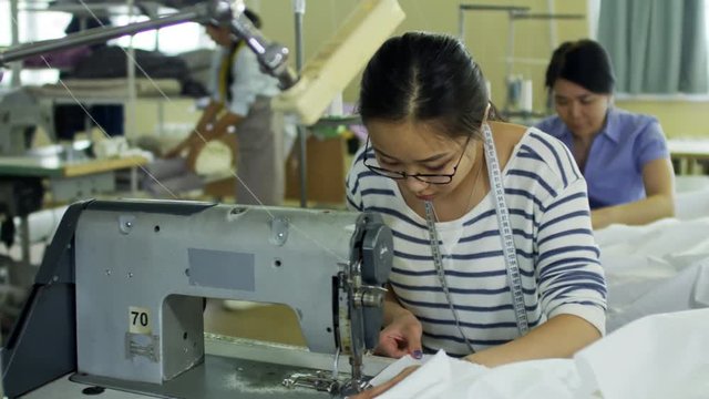 Medium shot of concentrated Asian seamstresses making clothes and using sewing machine at factory