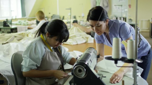 Young Hispanic woman in apron using sewing machine at factory, her Asian colleague guiding and helping her