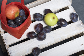 Ripe aromatic plums and apples in a wicker basket. Located on a wooden box, knocked out of the boards.