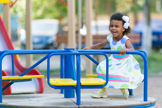 A black girl is riding a carousel. The child plays in the playground in the yard of the house.