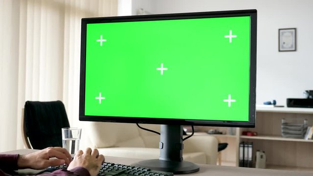 Unrecognisable man typing on personal computer keyboard with big green screen chroma mock-up in the living room of his house. Dolly slider 4K footage with parallax effect