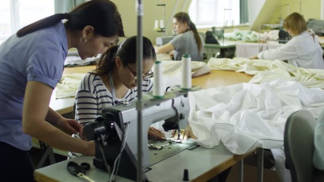 Young Asian Woman Trying To Make Clothes Using Sewing Machine, Her Colleague Guiding And Controlling Her