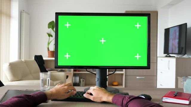 First person view - man hands typing on computer keyboard with big green screen chroma mock-up. The PC is on the desk in living room and the TV is on in the background