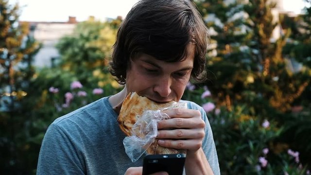 Dependence on the phone and the Internet. A man is using a smartphone and eats khachapuri - bread with cheese on the street, slow motion