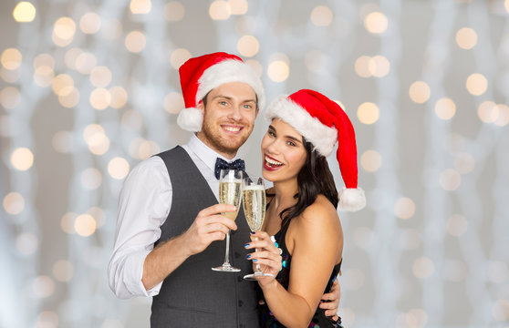 Celebration, People And Holidays Concept - Happy Couple In Santa Hats With Glasses Of Non Alcoholic Champagne At Christmas Or New Year Party Over Festive Lights Background