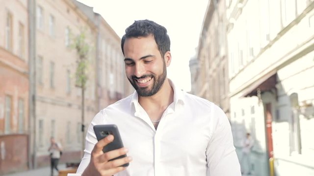 Smiling Man Using Mobile Phone And Walking Along City Street