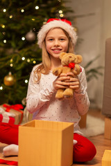 Obraz na płótnie Canvas christmas, holidays and childhood concept - smiling girl in santa helper hat with gift box and teddy bear at home
