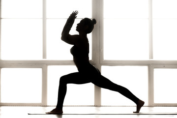 Fototapeta premium Young sporty attractive woman practicing yoga, doing Warrior one exercise, Virabhadrasana 1 pose, working out, wearing sportswear, indoor full length, at yoga studio, side view black silhouette