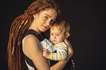 Portrait young happy mom with baby son in ergo backpack on dark wall on the background. Concept of modern parents