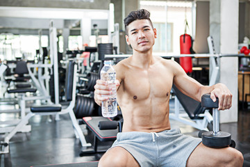 Fototapeta na wymiar Muscular bodybuilder guy sitting on a bench show water bottle and dumbbells after exercise in fitness gym . Shirtless young sportsman resting after training at the gym