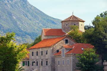 Fototapeta premium Red tiled roofs among green trees. Montenegro, Muo town. Residential buildings and ancient parish church (Church of the Blessed Virgin Mary)