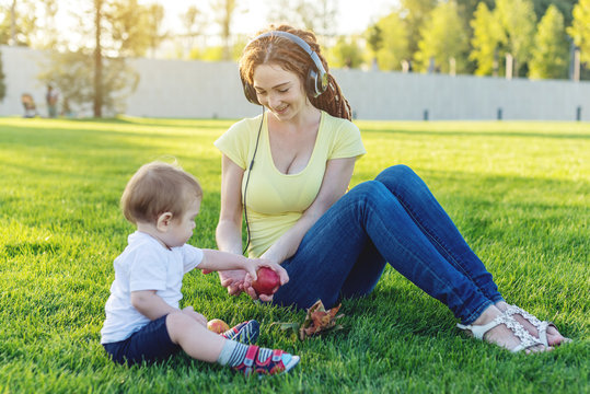 Young modern mom in headphones playing in a green meadow with her cute baby son in a Sunny Park. Joy of motherhood