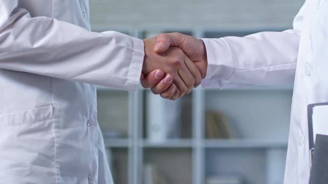Close up shot of two unrecognizable people in lab coats standing in office and shaking hands after reaching agreement