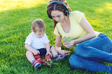 Naklejka na ściany i meble Young modern mom in headphones playing in a green meadow with her cute baby son in a Sunny Park. Joy of motherhood