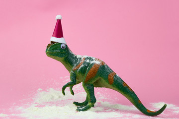 funny green dinosaur toy in little santa claus hat  and snow on pastel pink background