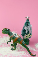funny green dinosaur toy near little christmas tree  on pastel pink background