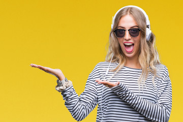 Beautiful young blonde woman wearing headphones and sunglasses over isolated background amazed and smiling to the camera while presenting with hand and pointing with finger.