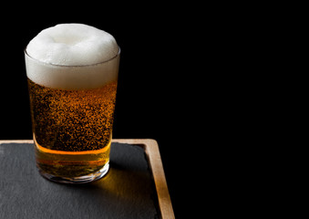 Glass of lager beer with foam and bubbles on stone board on black background. Space for text