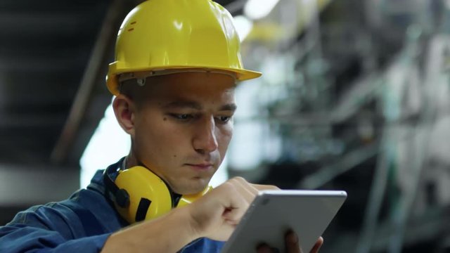 Close up face of concentrated male worker in bump cap and earmuffs using tablet computer when standing in industrial facility