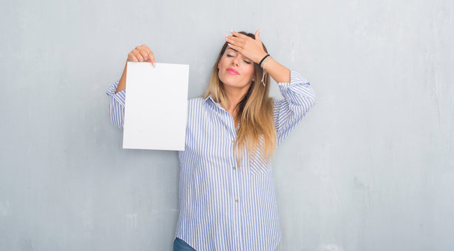 Young adult woman over grey grunge wall holding blank paper sheet stressed with hand on head, shocked with shame and surprise face, angry and frustrated. Fear and upset for mistake.