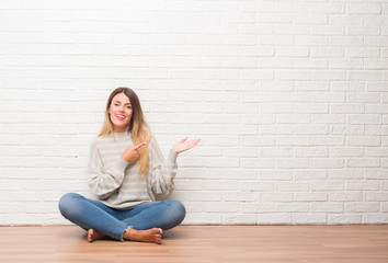 Young adult woman sitting on the floor over white brick wall at home amazed and smiling to the camera while presenting with hand and pointing with finger.