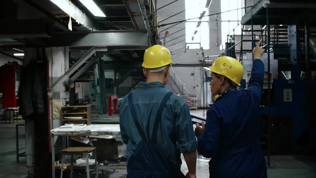 Rear view of male factory worker in yellow protective helmet and workwear listening to female colleague while walking through industrial building