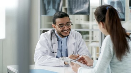 Tracking shot of smiling Asian doctor in lab coat and glasses sitting at desk in office and talking to female patient who is filling out form with information - Powered by Adobe