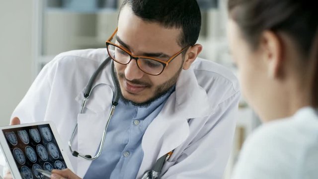 Medium shot of young Asian doctor in lab coat and glasses holding tablet computer and showing MRI pictures to unrecognizable female patient seen from her back