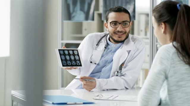 Tracking shot of friendly young Asian doctor sitting at desk in office and using laptop computer to show MRI pictures to female patient seen from her back