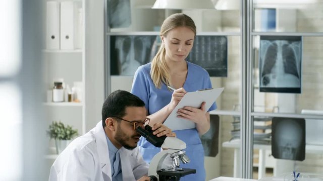 Tilt down shot of young black man looking into microscope in laboratory and discussing work with Caucasian colleague who is writing data into document attached to clipboard