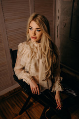Fototapeta na wymiar Beautiful luxurious blonde woman sitting on chair on a dark textural background. Studio shot. Girl looking outside